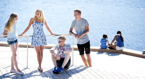 Två tjejer och två killar står och sitter tillsammans på en brygga. I bakgrunden sitter en mamma och ett barn och tittar ner i vattnet. 