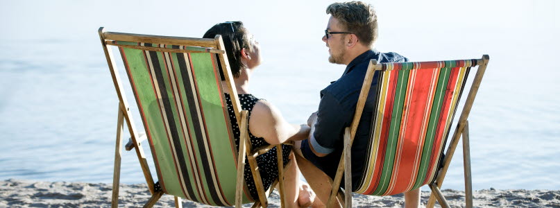 Man och kvinna sitter i varsin solstol på stranden vid vattnet